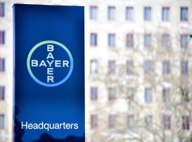 Primo piano di un totem col logo della Bayer davanti a un edificio con finesrte disposte a griglia