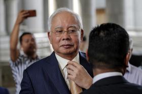Primo piano di Najib Razak che si sistema la cravatta; dietro di lui, sfocato, un uomo lo fotografa col cellulare