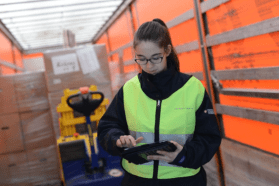 Donna che indossa una mantellina gialla inserisce dati su un tablet, circondata da merci di magazzino