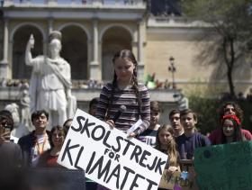 IGreta Thunberg in una piazza con giovani manifestanti si appresta a parlare a un microfono; attorno, monumenti e piante