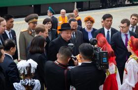 Un uomo con cappotto nero e cappello parla a dei giornalisti; dietro di lui, militare in uniforme, davanti donne in costume folk