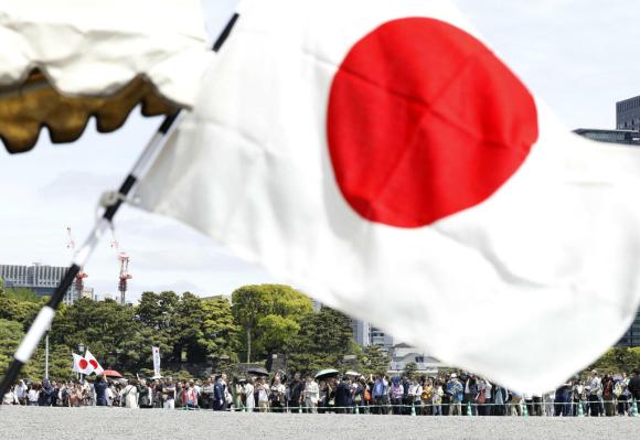 Primissimo piano di una bandiera giapponese. In lontananza, adunata di persone ai margini di un parco