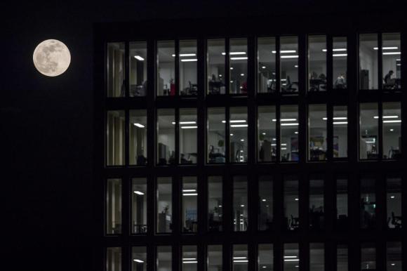 Luna piena ritratta alla sinistra di un palazzo di uffici con le luci interne accese, si intravvedono le scrivanie