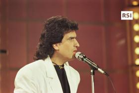 Toto Cutugno con maglietta nera e blazer bianco vicino a un asta microfono; si intravvedono palco e luci di scena
