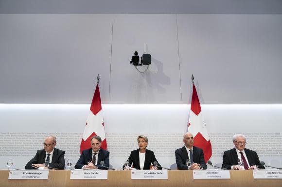 Banco di una sala conferenze con 5 persone in abito formale; allo loro spalle, due grosse bandiere svizzere e parete bianca