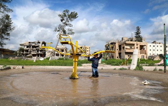 Un ragazzino spinge una giostra gialla in quel che resta d un parco giochi, nel fango e con edifici distrutti sullo sfondo