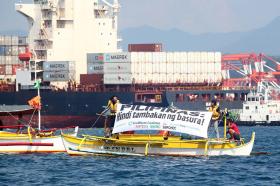 Immagine di una nave in mare aperto con una sessantina di container; più vicine, due barche con striscioni di protesta