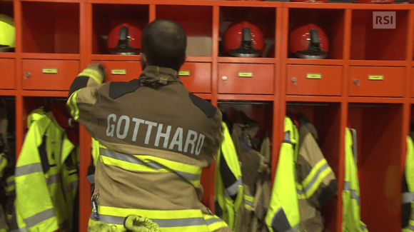 Pompiere visto di schiena (con tuta catarifrangente e scritta Gotthard ) mentre prende il suo casco da uno scaffale rosso