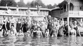 Gruppo di uomini e donne in acqua sul litorale di un lago; indossano costumi d altri tempi; costruzione in legno sul fondo