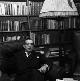 Uomo seduto su una poltrona, accanto a una lampada a stelo; dietro, libreria
