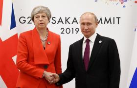 May in tailleur rosso e Putin in abito scuro si stringono la mano tra le bandiere britannica e russa; sul fondo logo del G20