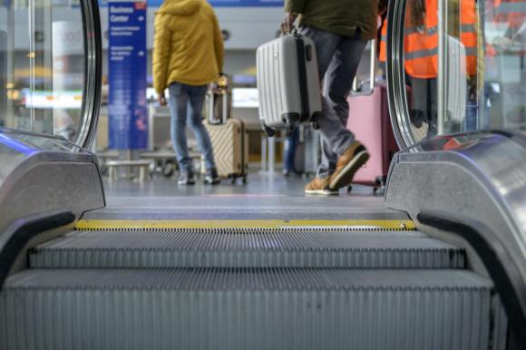 Parte finale di una scala mobile in ambiente aeroportuale; due persone col trolley e cartelli indicatori