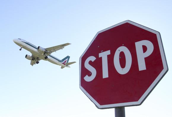 Primo piano di un cartello stradale Stop ; dietro, in cieli, un aereo con livrea di Alitalia sta prendendo quota