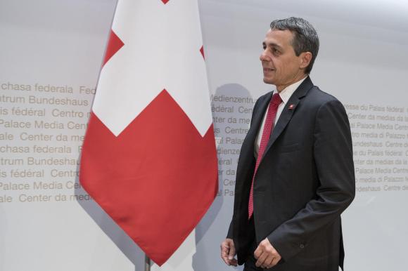 Uomo in abito formale con cravatta rossa, accanto a una bandiera svizzera; sul fondo, scritta Centro media Palazzo federale