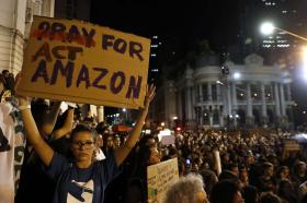 Piazza di città, di notte, colma di gente; in primo piano una ragazza con cartello Act for Amazon