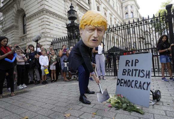 uomo travestito da Boris JOhnson con una pala davanti a un cancello e un cartello