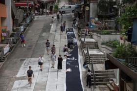 Telo nero steso lungo una strada cittadina lastricata in cemento; alcuni giovani vi dipingono con gessetti