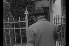 Uomo con cappotto e cappello, di schiena, varca il cancelletto di una palazzina numero civico 11