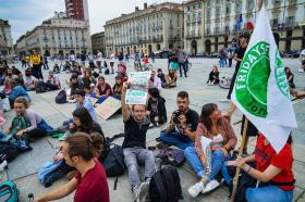 Gruppi di ragazzi seduti a gambe incrociate su una grande piazza cittadina, con cartelli ambientalisti