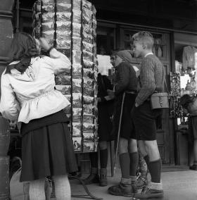 In un immagine in bianco e nero, girello espositore di cartoline con attorno alcuni ragazzini e ragazzine