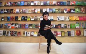 Donna vestita di nero sfoglia un libro, seduta su uno sgabello; dietro, mensole colme di libri, esposti