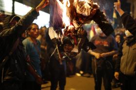 Esterno notte, gruppo di persone dà fuoco a un oggetto di tessuto con stampato un volto; manifestanti attorno