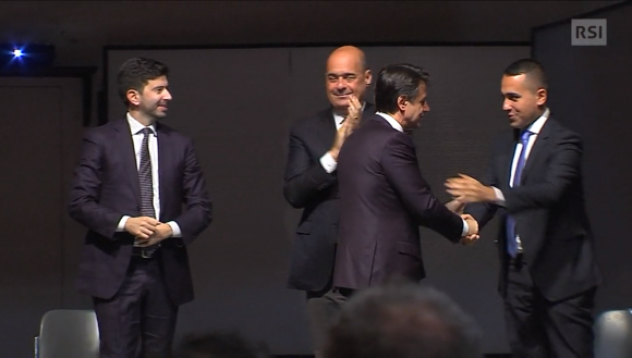 Quattro uomini in abito formale si stringono le mani, a turno, su un palcoscenico