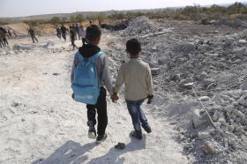 Due bambini, ritratti di schiena, camminano tenendosi per mano nei pressi di un cumulo di macerie in aperta campagna