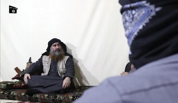 Uomo con abiti arabi e giberna da combattimento seduto a gambe incrociate con accanto un kalashnikov