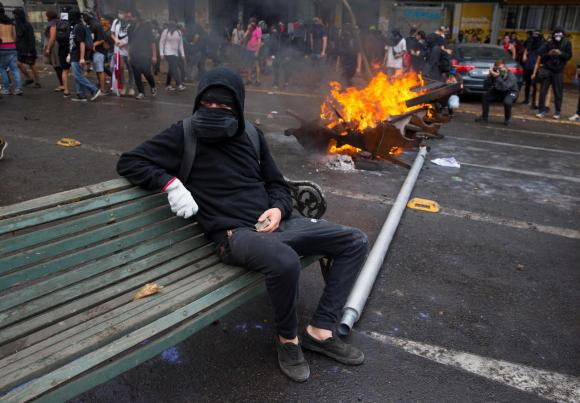 Un uomo completamente mascherato e vestito di nero seduto su una panchina mentre attorno le proteste impazzano