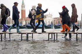 Turisti vestiti per freddo e pioggia camminano su una passerella elevata sopra l acqua; sul fondo, San Marco a Venezia