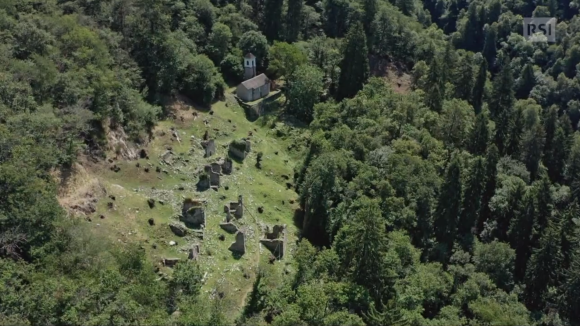 Vedura dall alta di una chiesetta immersa bei boschi; accanto, prato verde con rovine di rustici