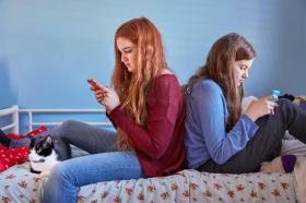 Due adolescenti, viste di profilo sedute su un letto, messaggiano con il telefonino