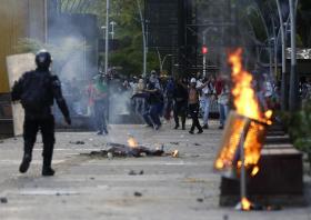 Strada con sassi, detriti e cestino della spazzatura in fiamme; manifestanti a volto coperto in lontananza; poliziotti di fronte