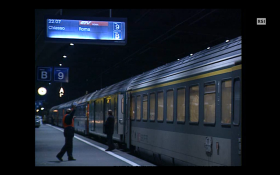 Banchina di stazione di notte; con treno e controllore e teleindicatore a palette con scritto EN Chiasso Roma