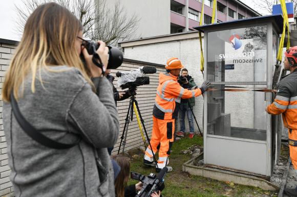 Due operai in tuta arancione assicurano una cabina telefonica a un argano per la rimozione; fotografi riprendono la scena