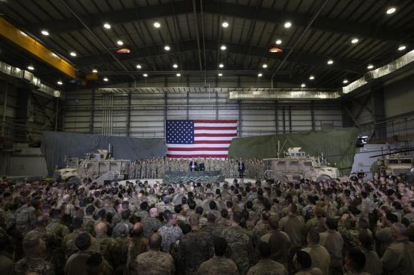 Interno di un capannone con, sul fondo, bandiera USA e presidente Trump al pulpito; davanti, centinaia di soldati