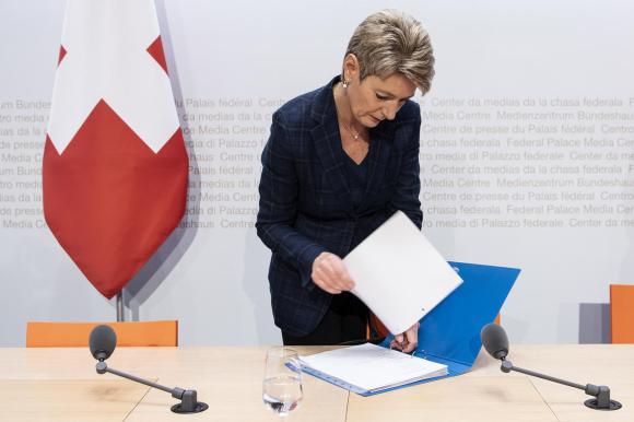 Donna in abito formale consulta un fascicolo su un tavolo con microfoni; dietro, bandiera svizzera e scritta Palazzo federale