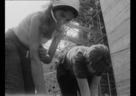 Due giovani, una ragazza e un ragazzo, intenti in un lavoro manuale. Sullo sfondo, un cantiere