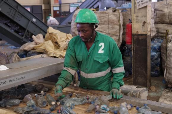 Un uomo che indossa tuta e guanti da lavoro controlla bottiglie di plastica usate su un nastro trasportatore industriale.