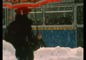 Un uomo con ombrello, ripreso di schiena, osserva il passaggio di un autobus in un paesaggio innevato.