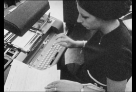 Giovane donna batte sulla tastiera di una macchina perforatrice i dati che copia da un foglio manoscritto.