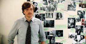 Un uomo in camicia e cravatta in piedi e sullo sfondo una parete su cui sono appese foto in bianco e nero di polizia.