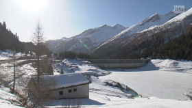 Lago artificiale, con diga di sbarramento ben in vista e superficie completamente ghiacciata; paesaggio alpino innevato