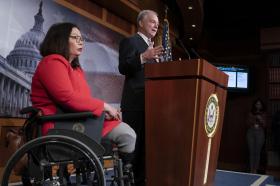 Un uomo in abito formale a un pulpito e una donna in abito rosso su sedia a rotelle accanto; dietro di essi immagine del Capitol