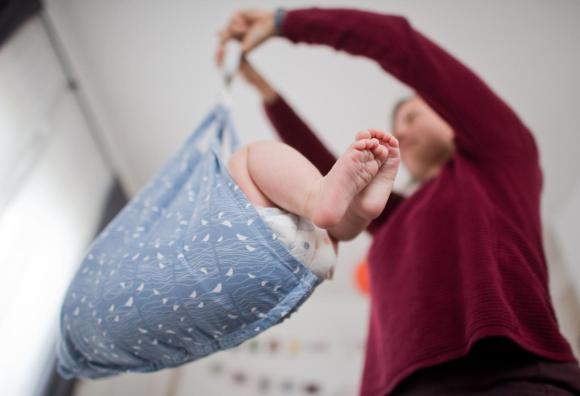 Signora vista dal basso (viso sfocato) pesa un bambino con una bilancia per neonati; spuntano le gambe del bebé