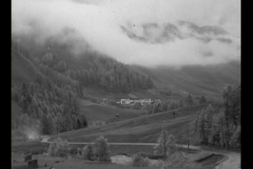 Foto in bianco e nero di unampia vallata alpina con boschi, campi e qualche nuvola