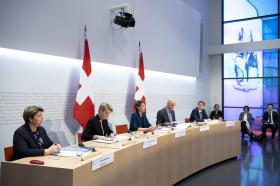 Sei persone sedute a un tavolo di conferenza stampa a semicerchio. Due bandiere svizzere dietro.
