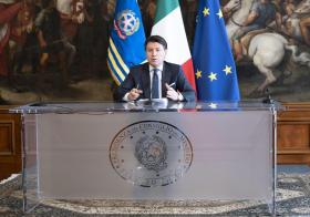 Uomo in abiti formali seduto a un pulpito con scritta Presidenza consiglio dei ministri Bandiera italiane ed europea dietro