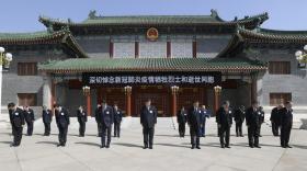 Gruppo di uomini in abito formale e scuro disposti in ranghi fanno un inchino; dietro, costruzione cinese